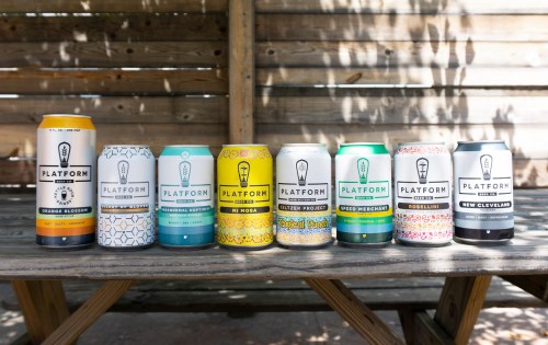 A lineup of colorful Platform beer cans on a wooden table against a wooden backdrop.
