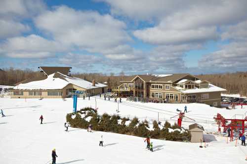 A snowy ski resort with a lodge, ski lifts, and people skiing on a clear day with clouds.