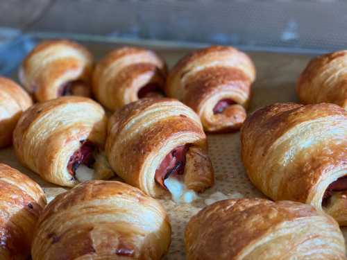 Freshly baked croissants with a golden-brown crust, some filled with cheese and ham, arranged on a baking tray.