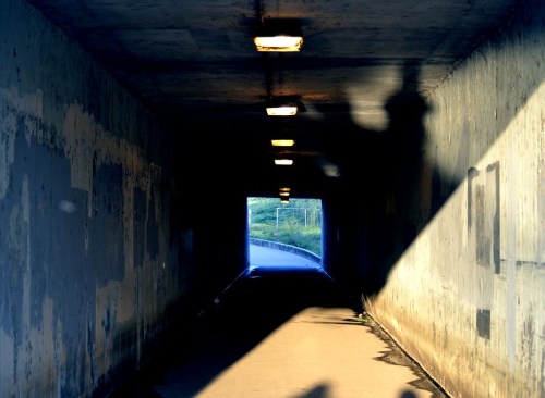 A dimly lit tunnel with concrete walls, leading to a bright exit, casting long shadows on the ground.