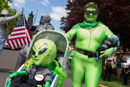 A woman in a green costume holds a doll and stands beside a stroller with an alien figure, while others celebrate in the background.