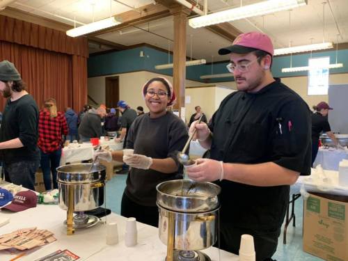Two volunteers serve food at a community event, smiling and working together at a table with cooking equipment.