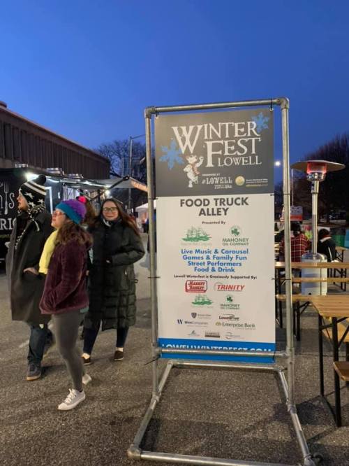 A sign for Winter Fest in Lowell, featuring food trucks, live music, and family fun, with people walking nearby.