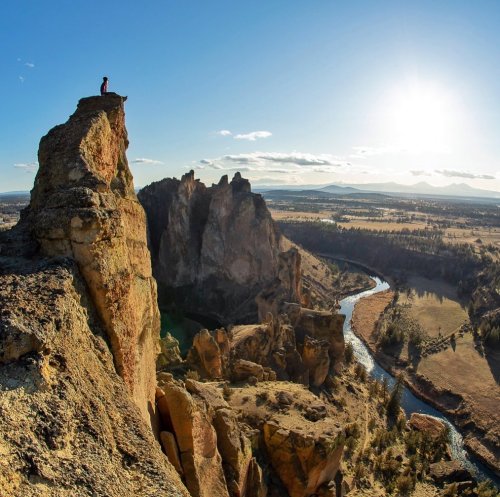 A person stands on a rocky peak overlooking a valley and river, with mountains and a bright sky in the background.