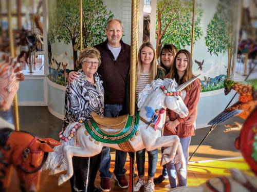 A family poses together in front of a carousel, with a unicorn and colorful animals in the background.