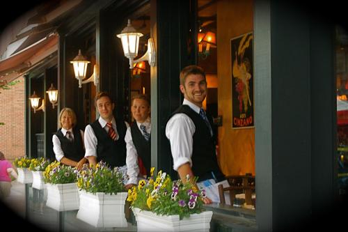 A group of four waitstaff in vests and ties smile from a restaurant window, surrounded by colorful flower boxes.