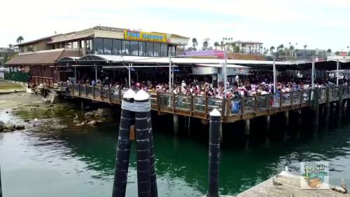 A busy waterfront restaurant with a large outdoor deck, filled with people, overlooking calm waters.