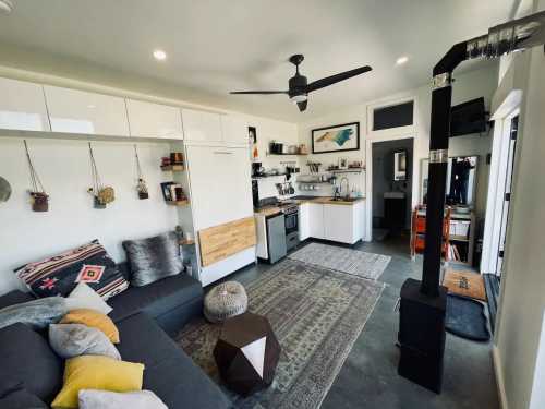 Cozy living space with a gray sofa, modern kitchen, decorative plants, and a wood stove, featuring warm lighting and stylish decor.