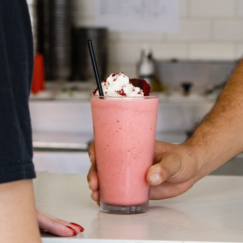 A hand holds a pink smoothie topped with whipped cream, while another hand reaches out to it.