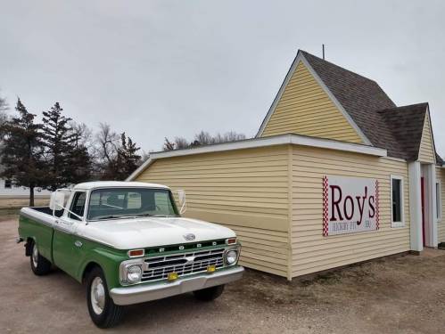 A vintage green and white Ford truck parked outside a yellow building with a sign that reads "Roy's."