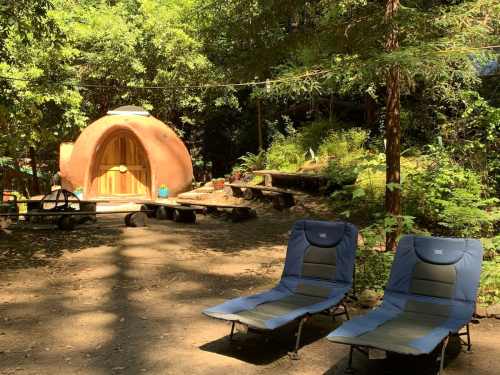 A cozy, round clay structure surrounded by greenery, with two blue lounge chairs in the foreground.