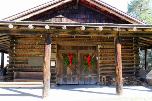 A rustic log cabin with wooden pillars, featuring festive wreaths with red bows on the doors.