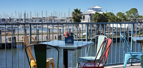 Colorful chairs at a waterfront restaurant with a view of boats and a marina under a clear blue sky.