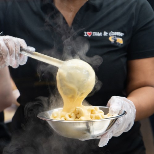 A person in a black shirt holds a bowl of steaming mac and cheese, with cheese stretching from the bowl.