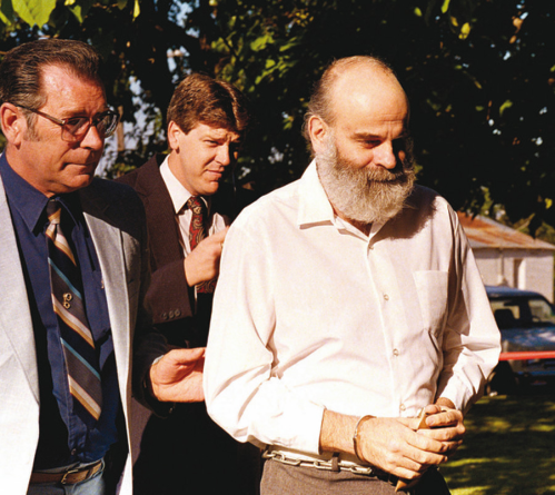 Two men escort a bearded man in handcuffs outdoors, with trees and a building in the background.