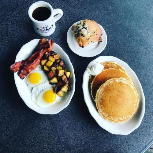 A breakfast spread featuring eggs, bacon, potatoes, pancakes, and a blueberry muffin, with a cup of coffee.