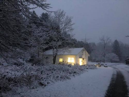 A snowy landscape at dusk, featuring a cozy house with warm light glowing from its windows. Trees are covered in snow.