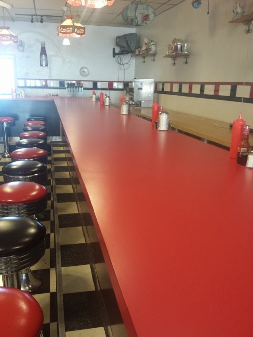 A retro diner interior featuring a long red counter with black and red stools, condiments, and a checkered floor.