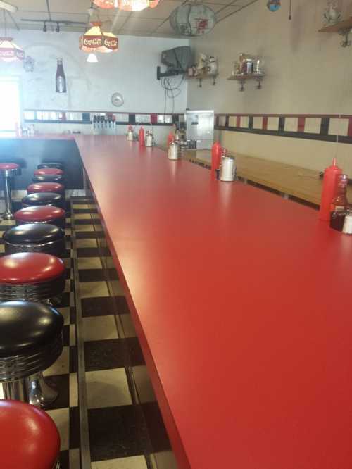 A retro diner interior featuring a long red counter with black and red stools, condiments, and a checkered floor.