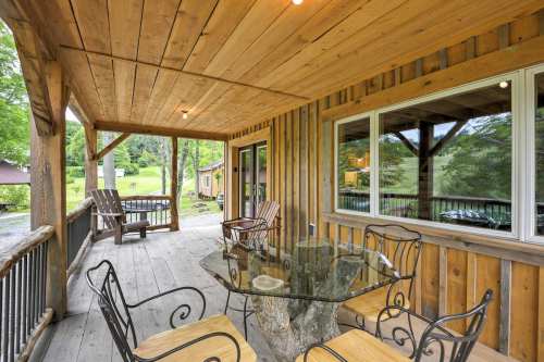 A wooden porch with a glass table and chairs, surrounded by greenery and rustic cabins in the background.