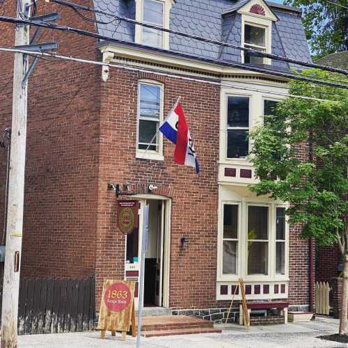 A brick building with a sign reading "1863" and flags displayed, surrounded by greenery and power lines.