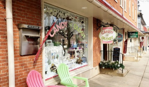 A storefront with a colorful candy shop window display and two green chairs outside on a brick sidewalk.
