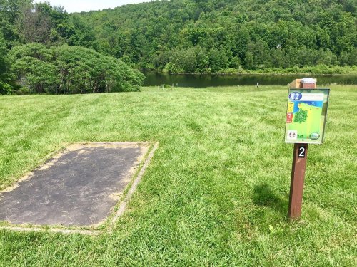 A grassy area with a disc golf tee pad and a sign for hole 12, overlooking a river and wooded hills in the background.