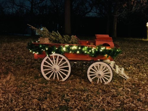 A festive red wagon decorated with greenery and lights, set against a dark, leaf-covered landscape.