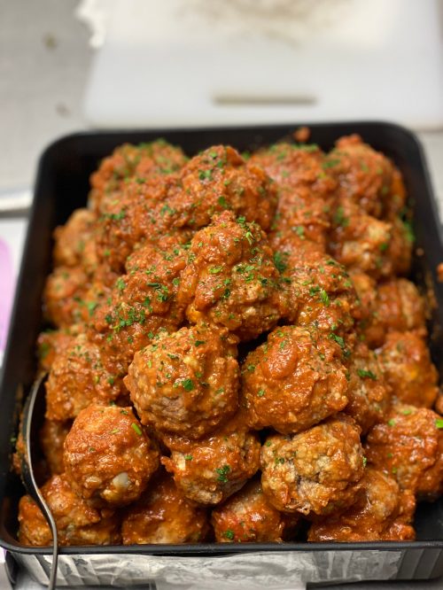 A large tray filled with golden-brown meatballs coated in a rich red sauce and garnished with chopped parsley.