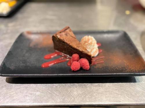 A slice of chocolate cake on a black plate, garnished with raspberry sauce and fresh raspberries.