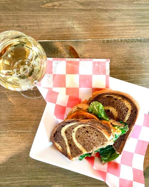 A sandwich with leafy greens on dark bread, served on a white plate with a glass of white wine beside it.