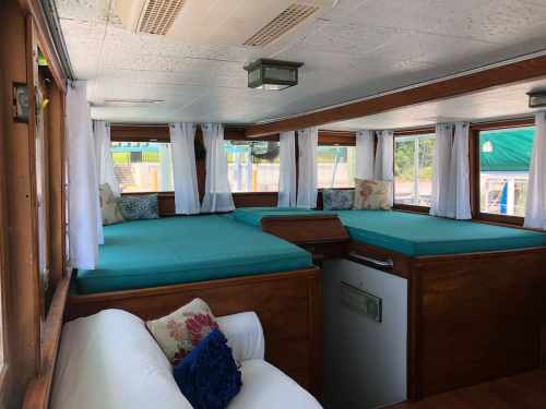 Interior of a boat featuring two large beds with teal mattresses, white curtains, and a cozy seating area.