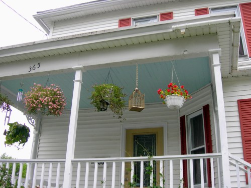 A white house porch with hanging flower baskets and a birdcage, featuring red shutters and a welcoming entrance.