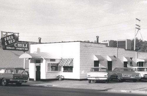 Black and white photo of a vintage diner with striped awnings and classic cars parked outside. Sign reads "Fred & Red Chili."