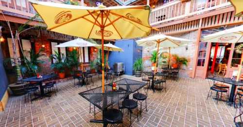 Outdoor dining area with metal tables, chairs, and yellow umbrellas surrounded by greenery and brick walls.