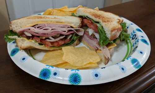 A plate with a toasted sandwich filled with ham, lettuce, tomato, and onion, accompanied by tortilla chips.