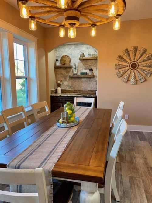 A rustic dining room with a wooden table, white chairs, and a chandelier, featuring a cozy kitchen nook in the background.
