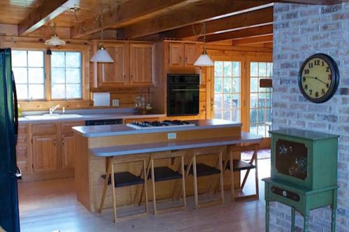 A cozy wooden kitchen with a central island, three stools, and a vintage clock on the wall. Natural light fills the space.