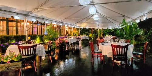 A beautifully set outdoor dining area under a tent, featuring tables with white linens and ambient lighting.