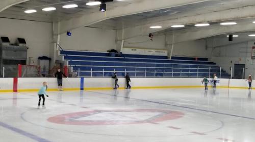 A group of children ice skating on a rink with empty bleachers in the background.