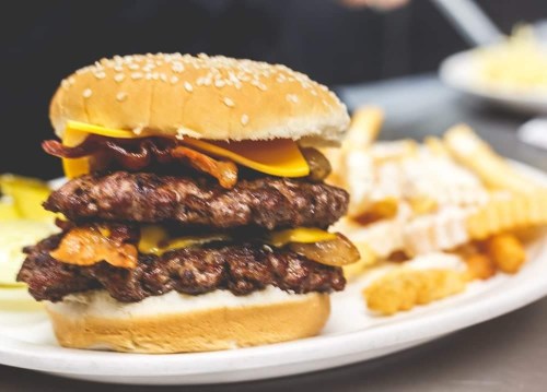 A stacked burger with two patties, cheese, bacon, and pickles, served with crinkle-cut fries on a plate.