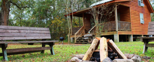 A cozy wooden cabin surrounded by trees, with a fire pit and benches in the foreground.