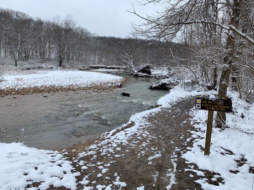 A snowy riverbank with a winding stream and a sign for a nature trail in a winter landscape.