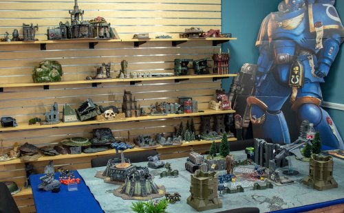A tabletop gaming setup featuring detailed terrain and miniatures, with a backdrop of a large character cutout.