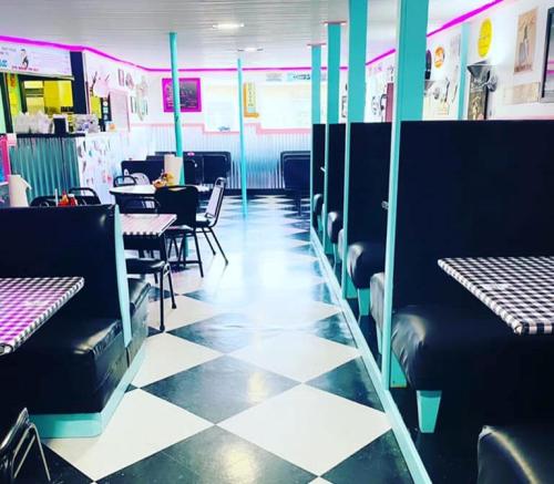 A retro diner interior with black and white checkered floors, teal accents, and cozy booths under pink neon lights.