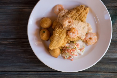 A plate of fried fish topped with shrimp and creamy sauce, served with coleslaw and golden corn fritters.