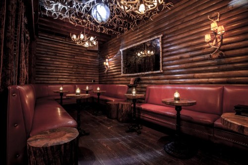 Cozy, rustic lounge with wooden walls, leather seating, candlelit tables, and antler decor.