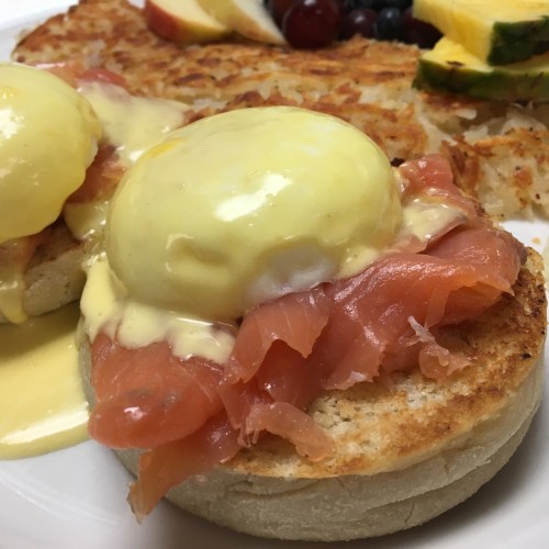Two poached eggs on toasted English muffins topped with smoked salmon and hollandaise sauce, served with fruit.