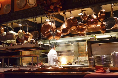 A cozy restaurant kitchen with hanging copper pots and a chef cooking over an open flame. Warm, inviting atmosphere.