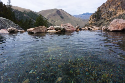 A serene natural hot spring surrounded by rocky hills and clear water, reflecting the landscape above.